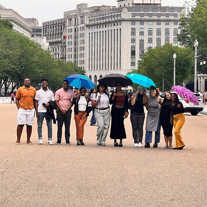 10 LAF Ignite students pose smiling on a busy pedestrian street with some holding umbrellas