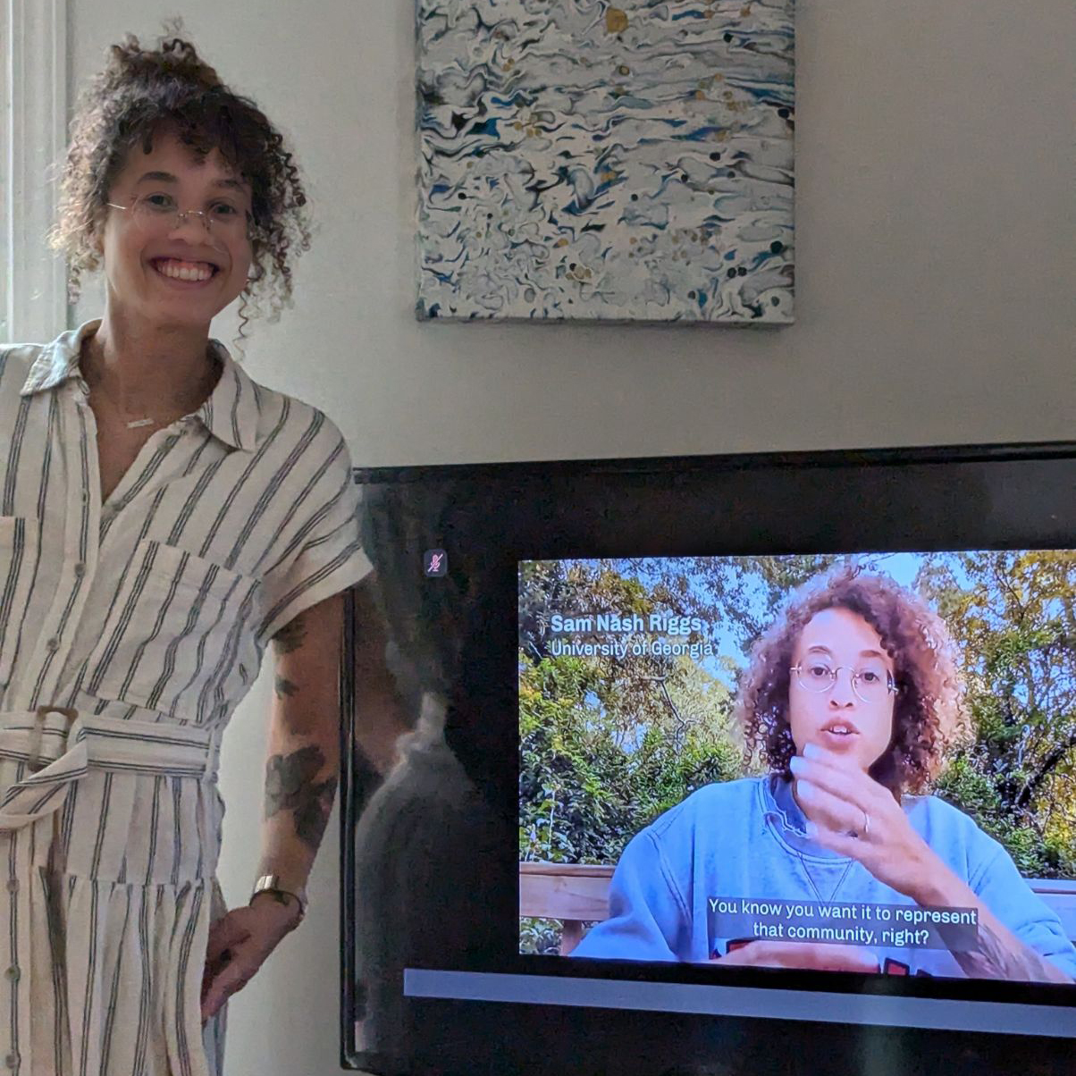 Sam Nash Riggs smiling and standing in front of a television which is showing her recorded clip from the Olmsted Scholar Virtual Induction ceremony.