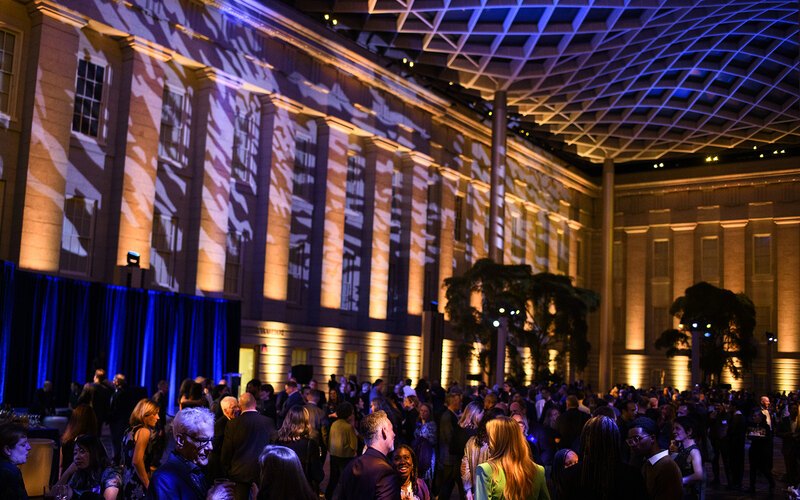 Blue lights and shadows appear on the undulating glass ceiling and historic building facade above the crowd of Annual Benefit attendees in the Kogod Courtyard