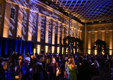 Blue lights and shadows appear on the undulating glass ceiling and historic building facade above the crowd of Annual Benefit attendees in the Kogod Courtyard