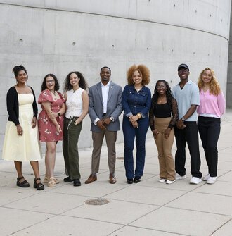 A group of 8 LAF Ignite students stand in front of a curved concrete wall.