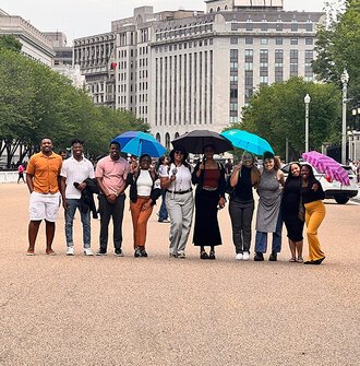 10 LAF Ignite students pose smiling on a busy pedestrian street with some holding umbrellas
