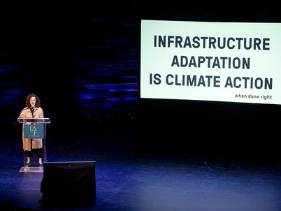 A woman stands at a podium on a dark stage with a large screen behind them. The screen displays the text 'INFRASTRUCTURE ADAPTATION IS CLIMATE ACTION when done right.'