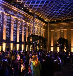 Blue lights and shadows appear on the undulating glass ceiling and historic building facade above the crowd of Annual Benefit attendees in the Kogod Courtyard